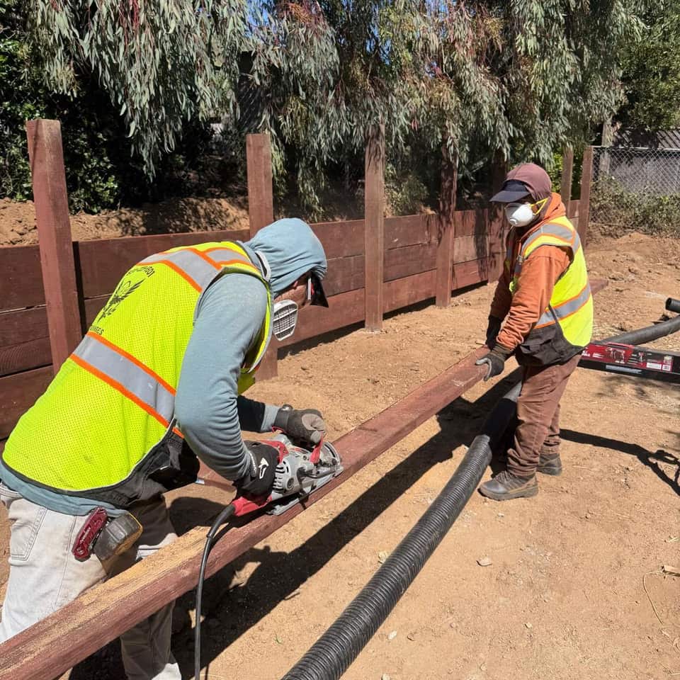Two crew members cutting and fitting a redwood fence rail on a hillside lot